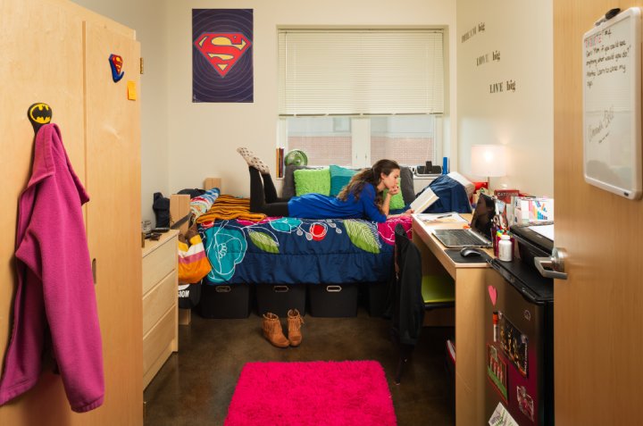A student lies on a bed in a dorm room, reading a book. The room is decorated with a colorful bedspread, Superman poster, motivational quotes, and a pink rug. A desk is organized with books and a laptop, while storage bins sit under the bed.